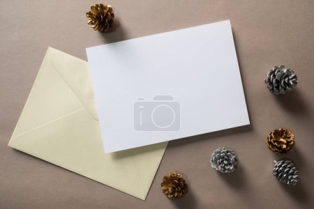 Photo for Christmas winter festive ornament. Blank paper card and envelope with silver and golden pine cones on brown background. Minimal decoration. Invitation Christmas holiday party. Flat lay, top view, copy space. - Royalty Free Image