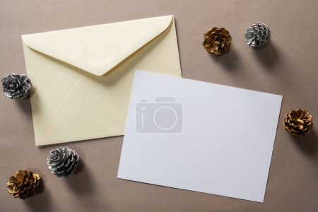 Photo for Christmas winter festive ornament. Blank paper card and envelope with silver and golden pine cones on brown background. Minimal decoration. Invitation Christmas holiday party. Flat lay, top view, copy space. - Royalty Free Image