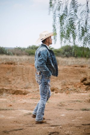 A cowboy wearing jeans and a hat, standing under the sky with fluffy clouds, working on a farm
