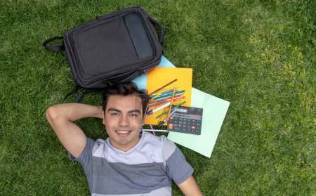 Male student lying on the grass with a backpack and school supplies