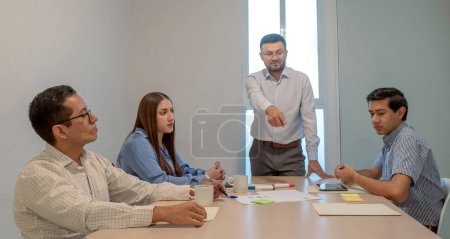 Mann zeigt auf einen Mann vor einer Gruppe von Menschen, die um einen Tisch sitzen