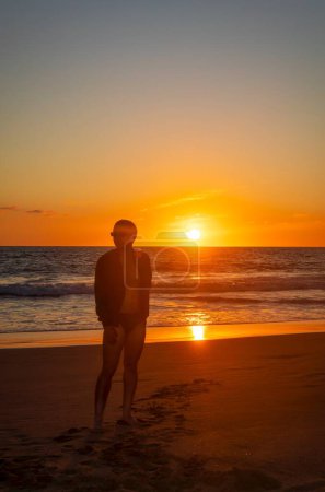 Téléchargez les photos : Un jeune homme debout sur la plage profitant d'un coucher de soleil ensoleillé au bord de la mer - en image libre de droit