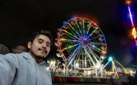 A Man taking selfie at Mexican fair with ferris wheel and colorful lights at night