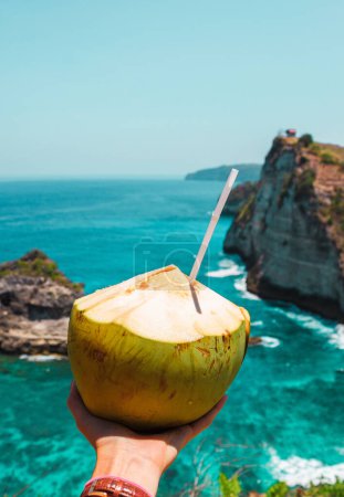 Photo for A hand with fresh coconut on the ocean and cliffs tropical background. Tropical landscape. Vacation mood. Bali, Indonesia - Royalty Free Image