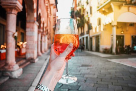 Photo for Female hand with glass of orange cocktail spritz on the background of old buildings. Alcoholic drink and ice. Sunny summer day in Verona, Italy. Vacation in Europe. - Royalty Free Image