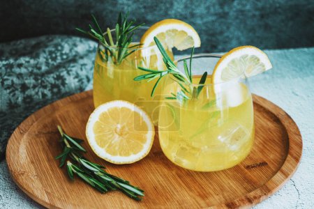 Photo for Glasses with cold cocktail, lemonade, sliced lemon and rosemary plant on the concrete table. Wooden plate, drink close up still life. High quality photo - Royalty Free Image