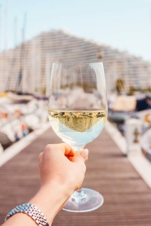 Photo for Woman hand with white wine, champagne glass on a yachts, sailing boats, building background. Vacation in Europe. Nice, French riviera, bay. Drink photo. Romantic relax, holiday on the sea coast. - Royalty Free Image