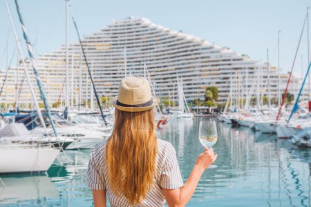 Photo for Blonde hair woman in a hat standing with her back on sea, yachts, buildings and boats background. White wine glass in hand. Vacation in Europe. Nice, French riviera. Travel photo. - Royalty Free Image