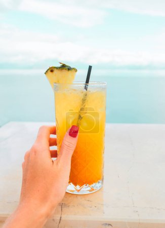 Photo for Female hand with a red manicure holds a tropical yellow-orange cocktail on the background of the sea. Vacation photo. High quality photo - Royalty Free Image
