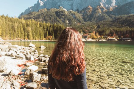 Photo for A young woman with long brown hair stands on the background of a lake Eibsee in the mountains of the German Alps. Autumn walk in Bavaria. - Royalty Free Image