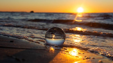 Photo for A glass ball lies in the waves on the sandy beach, the sea and the setting sun are reflected in the ball - Royalty Free Image