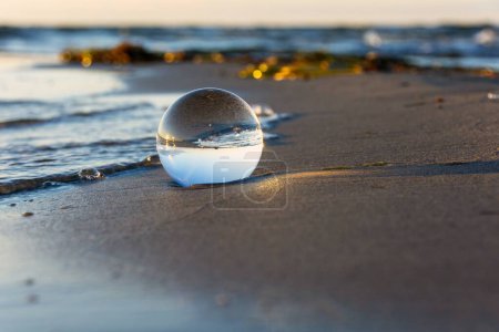 Une boule de verre se trouve dans les vagues sur la plage de sable, la mer et le soleil couchant se reflètent dans la boule