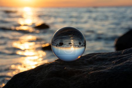 Glaskugel auf einem Felsen bei Sonnenuntergang am Strand, das Meer und die untergehende Sonne spiegeln sich in der Kugel