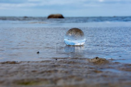 A glass ball lies in the waves on the sandy beach, the sea and the setting sun are reflected in the ball