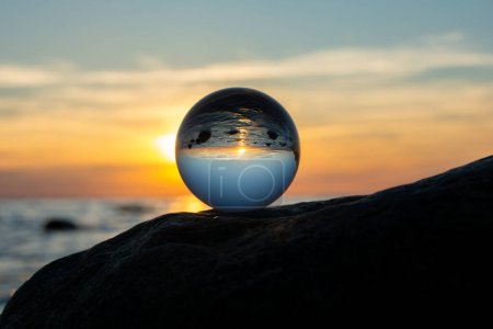 Glass ball on a rock at sunset on the beach, the sea and the setting sun are reflected in the ball