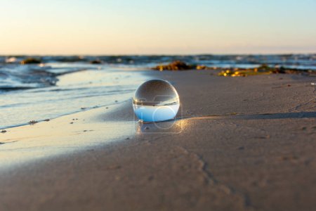 Eine Glaskugel liegt in den Wellen am Sandstrand, das Meer und die untergehende Sonne spiegeln sich in der Kugel