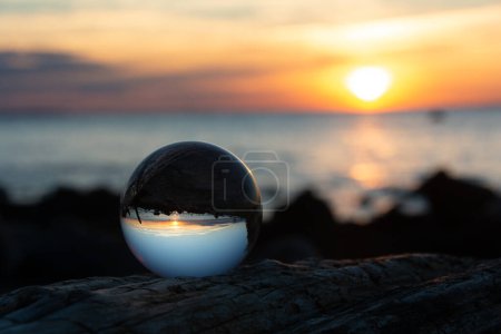 Glaskugel auf einem Ast bei Sonnenuntergang am Strand, das Meer und die untergehende Sonne spiegeln sich in der Kugel