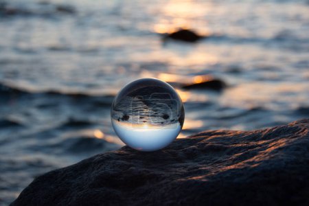 Glaskugel auf einem Felsen bei Sonnenuntergang am Strand, das Meer und die untergehende Sonne spiegeln sich in der Kugel
