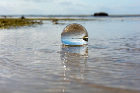Eine Glaskugel liegt in den Wellen am Sandstrand, das Meer und die untergehende Sonne spiegeln sich in der Kugel