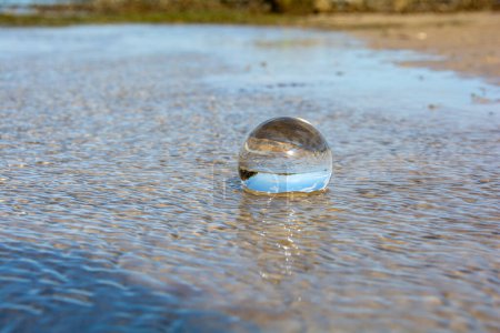 Una bola de vidrio se encuentra en las olas en la playa de arena, el mar y el sol poniente se reflejan en la pelota