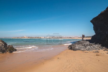 Téléchargez les photos : Homme âgé avec un sac à dos debout sur un rocher profitant de la vue sur Praia das Furnas dans la région d'Odemira, dans l'ouest du Portugal. Errant le long du sentier des Pêcheurs, Rota Vicentina. - en image libre de droit