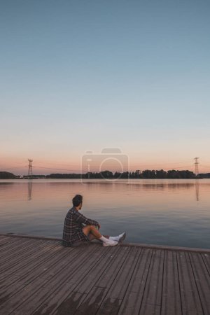 Photo for Young 25 year old brown man with plaid shirt sitting on the end of a wooden pier during sunset, watching the calm water surface in Almere Netherlands. - Royalty Free Image