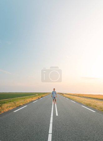 Photo for Brown-haired man in a checked shirt walks along a road at sunset on the outskirts of Almere, Netherlands. Fashion lifestyle. - Royalty Free Image