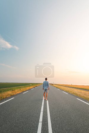 Photo for Brown-haired man in a checked shirt walks along a road at sunset on the outskirts of Almere, Netherlands. Fashion lifestyle. - Royalty Free Image