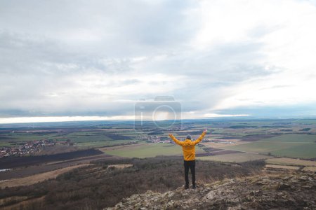 Traveller in a yellow jacket stands on top of Mount Rip, raising his arms above his head. Achieving success. Exploring the Czech land.