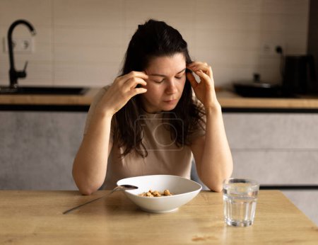 traurige junge Frau, die ihren Kopf mit den Händen hält, Frühstück, einen Teller Haferflocken und ein Glas Wasser