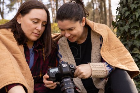 two young women in the forest under a blanket are looking at a photo on a camera