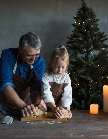 Foto de Papá y su hija amasan la masa juntos en la mesa y preparan golosinas de Navidad. Las tradiciones familiares - Imagen libre de derechos