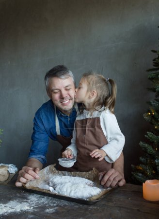 Foto de Una pequeña hija besa a su padre en la mejilla mientras cocina un regalo de Navidad en la cocina. - Imagen libre de derechos