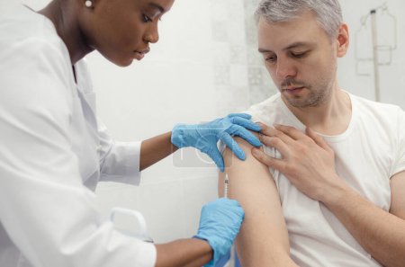 Téléchargez les photos : An Afro-American nurse vaccinates a patient with a syringe in a medical office. - en image libre de droit