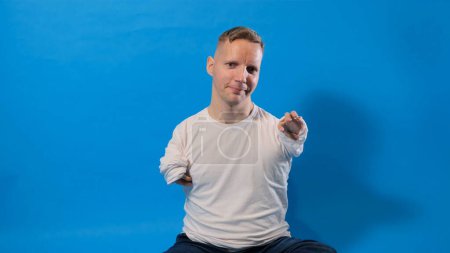 Foto de Hola Retrato de un hombre amigable y alegre con discapacidades en una camiseta blanca casual, mostrando un gesto de bienvenida, agitando su mano y sonriendo sinceramente sobre un fondo azul. - Imagen libre de derechos