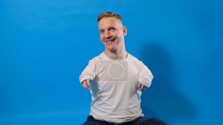Téléchargez les photos : Un homme souriant et joyeux dans un T-shirt blanc avec des handicaps assis sur un fond bleu isolé - en image libre de droit