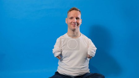 Foto de Un hombre sonriente y alegre en una camiseta blanca con discapacidades se sienta sobre un fondo azul aislado - Imagen libre de derechos