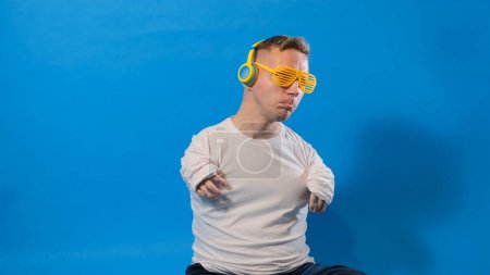 Téléchargez les photos : Un homme handicapé dans des lunettes élégantes et écouteurs jaunes écoute de la musique et danse sur un fond bleu dans le studio - en image libre de droit