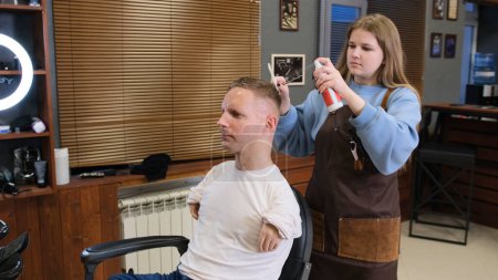 Foto de El maestro de la chica en la peluquería lleva a cabo el peinado del cabello para un hombre con discapacidad sentado en una silla de barbero - Imagen libre de derechos