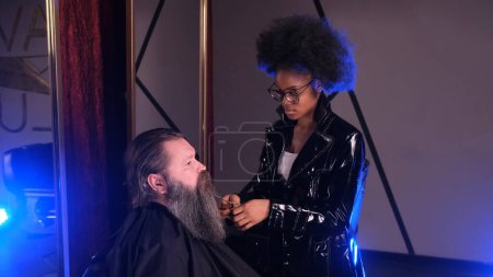 Photo for An African-American girl in a barbershop braids pigtails into the beard of a male client. - Royalty Free Image