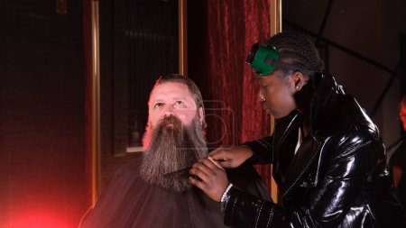 Foto de Una chica afroamericana en una barbería trenzas coletas en la barba de un cliente masculino. - Imagen libre de derechos