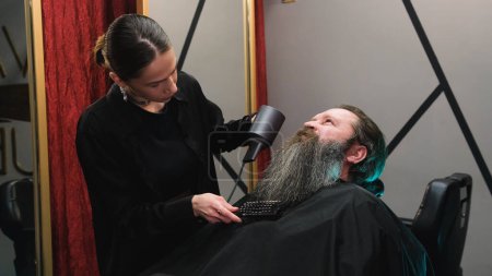 Téléchargez les photos : Une coiffeuse utilisant un sèche-cheveux pour sécher la longue barbe d'un homme dans un salon de coiffure. - en image libre de droit