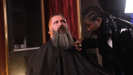 Téléchargez les photos : Une styliste afro-américaine nivelle la barbe d'un homme avec des ciseaux dans un salon de coiffure. - en image libre de droit