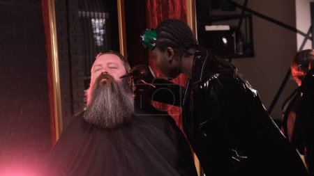 Foto de Una estilista afroamericana con un impermeable de cuero negro peina la barba de un cliente en una peluquería - Imagen libre de derechos
