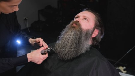 Téléchargez les photos : Une coiffeuse coupe la barbe d'un homme avec une tondeuse - en image libre de droit