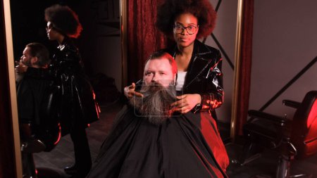 Foto de Una peluquera afroamericana endereza la barba con las manos a un cliente masculino sentado en un sillón - Imagen libre de derechos