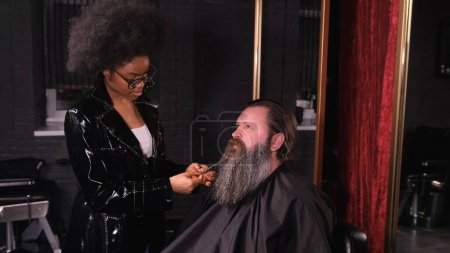 Foto de Una chica afroamericana en una barbería trenzas coletas en la barba de un cliente masculino. - Imagen libre de derechos