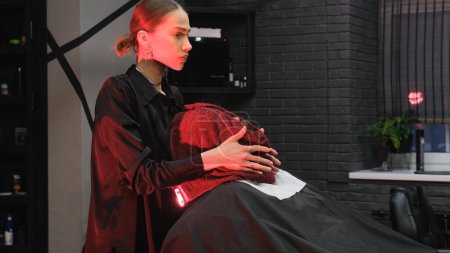 Foto de El barbero cubre la cara del hombre con una toalla caliente. Un cliente con una toalla caliente en la cara antes de afeitarse en una peluquería - Imagen libre de derechos