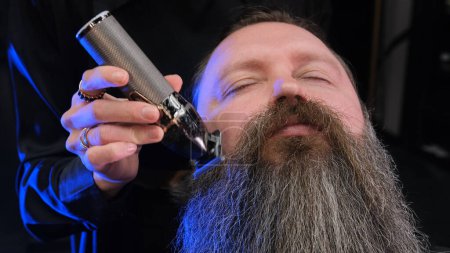 Foto de Un barbero corta la barba de un hombre con una máquina de escribir eléctrica de cerca - Imagen libre de derechos