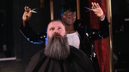 Téléchargez les photos : Une coiffeuse américaine tient des ciseaux dans ses mains, regarde la caméra et sourit. Un homme avec une grosse barbe est assis dans un fauteuil de coiffeur - en image libre de droit